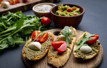 Bruschettas with pesto, tomatoes, mozzarella cheese and basil on wooden board served with guacamole, arugula and salt with herb. Italian tosted bread with vegetables and traditional sauce