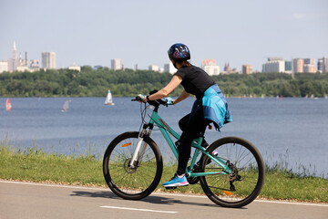 Girl in sportswear and helmet riding bicycle on river and city background. Cycling and summer leisure
