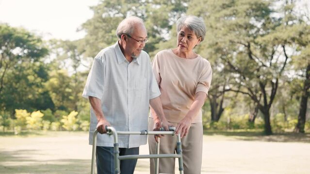 Asian senior woman prop up and consolation husband walking with walker cane in nature park, physical therapy concept