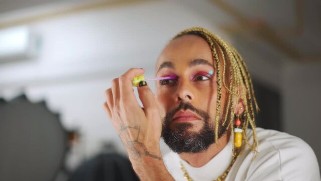 Male Caucasian extravagant transsexual man applying mascara on eyelashes. Close-up portrait of confident male queer doing makeup standing near mirror. Cosmetics and gender identity