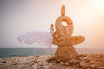 Woman sky stone. Lady stands on stone sculpture with ocean view. She is dressed in a white long...