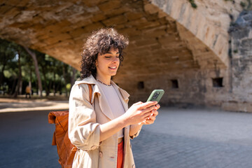 Young happy smiling beautiful female tourist using online maps on smart mobile phone to find city attractions while traveling, standing outdoors on sunny day.