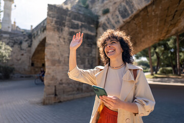Fototapeta premium Young woman with mobile phone waiving hand, saying hello on the street, happy and smiling, friendly welcome gesture.
