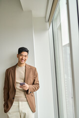 Happy Asian business man using mobile cell phone technology standing in office at window. Young...