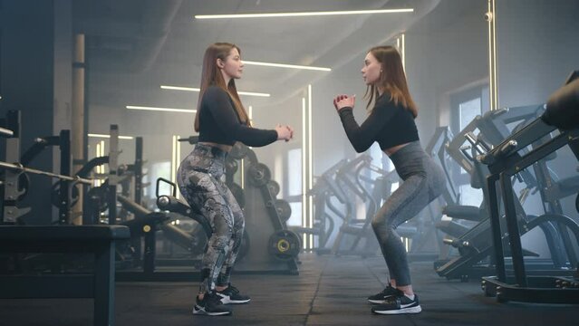 Profile two shot of young fit pretty brown-haired women performing squats in gym. Women stand opposite each other, look at each other and perform squats. High quality 4k footage