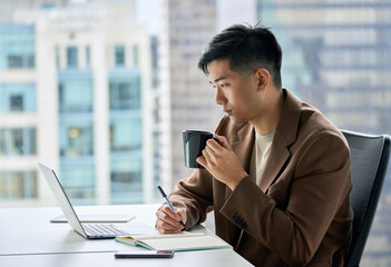 Young busy Asian business man employee working on laptop in corporate office writing notes. Professional businessman drinking coffee using computer, watching webinar, elearning sitting at desk.