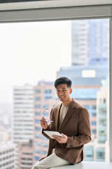 Fototapeta premium Young happy Asian business man professional manager, smiling Japanese businessman wearing suit using digital tablet sitting in corporate modern office with big window, vertical portrait.