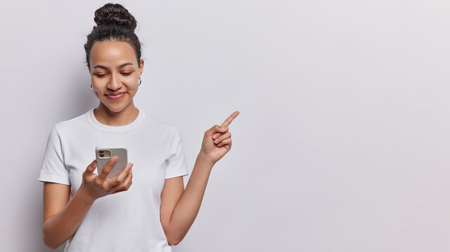 Stunning Latin woman with phone points at copy space showcases digital content and advertising messages promots products smiles pleasantly dressed in casual t shirt isolated over white background