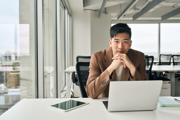 Young serious Asian businessman corporate manager looking at camera thinking working on laptop in...