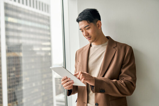 Young adult elegant professional Asian business man manager investor holding tablet using digital pc standing at office window doing digital data research on tech website analyzing financial market.