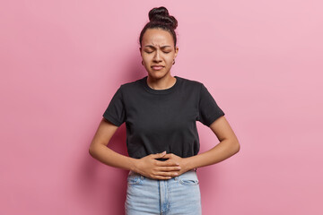 Upset sad worried Latin woman has stomach ache disorder feels unwell suffers from indigestion feels abdominal pain or cramps dressed casually isolated over pink background. Health problems concept