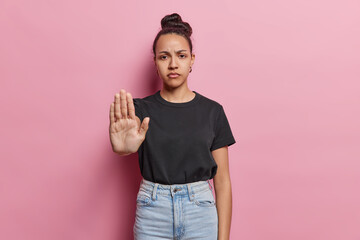 Serious Latin woman shows stop gesture with palm asserts boundaries sends message of resistance has strict expression wears black t shirt and jeans poses against pink background. Self empowerment - obrazy, fototapety, plakaty