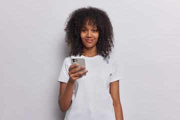 Pleased curly haired woman with satisfied expression holds mobile phone types message or scrolls news in internet dressed in casual t shirt isolated on white background. People and technology concept
