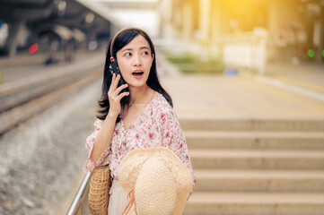 Fototapeta premium Asian young woman traveler with weaving hat using a mobile phone beside railway train station in Bangkok. Journey trip lifestyle, world travel explorer or Asia summer tourism concept.