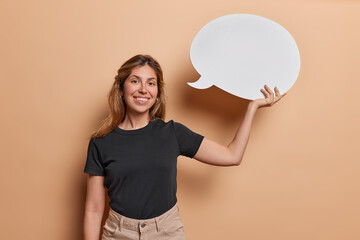 Horizontal shot of pleased woman symbolizes invitation for your promotional content or message holds white blank chat bubble smiles toothily wears black t shirt isolated over brown background