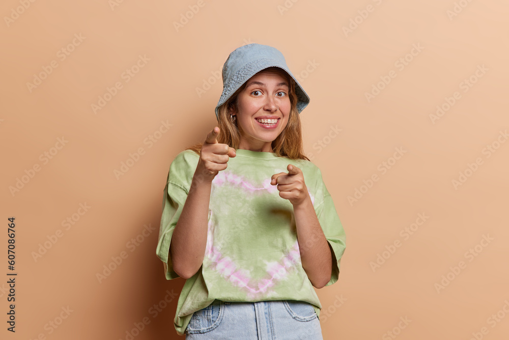 Wall mural smiling young pretty woman joyfully extends her index fingers towards camera selects someone with be