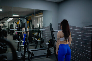 A sports girl in blue leggings and a sexy body warms up in front of a mirror at a workout in a gym with exercise equipment	