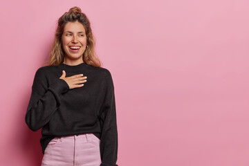 Horizontal shot of European woman holds hands on chest and smiles from happiness being fascinated dressed in casual black jumper and trousers isolated over pink background copy space for your text