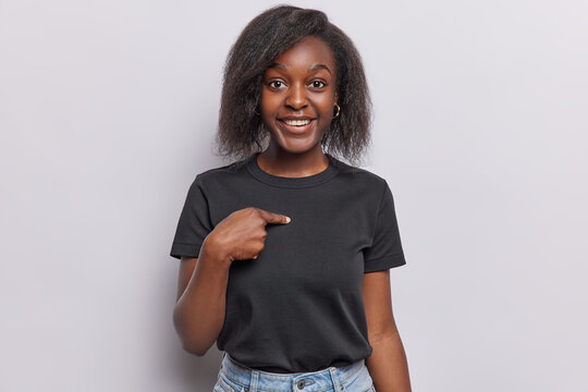 Surprised cheerful Afro woman points at herself asks who me dressed in casual black t shirt smiles pleasantly isolated over white background. Lovely dark skinned female model being chosen or picked up
