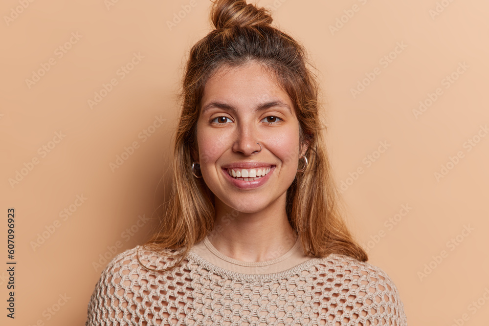 Poster Portrait of cheerful young European woman without makeup looks gladfully at camera smiles broadly dressed in knitted jumper wears earrings and piercing in nose isolated over brown background.