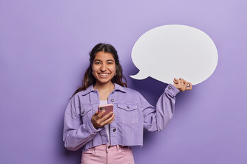 Happy brunette near east woman browses internet on mobile phone holds white communication bubble wears cropped denim jacket and pink trousers isolated over purple background. Technology concept