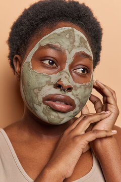 Thoughtful chubby African woman with short dark hair applies nourishing clay mask undergoes anti aging procedures touches cheek gently isolated over brown background. Beauty treatment concept