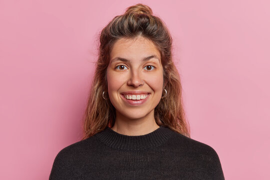 Portrait of cheerful European woman smiles broadly shows white perfect teeth glad to pose for making photo dressed in black jumper isolated over pink background. Positive human emotions concept