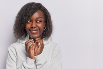 Horizontal shot of dark skinned woman steeples fingers has evil intention to do somethung thinks over scheme looks with cunning expression dressed in casual hoodie isolated over white background - obrazy, fototapety, plakaty