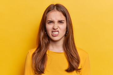 Portrait of dark haired young European woman keeps teeth clenched frowns face being irritated dressed in casual jumper isolated over vivid yellow background. People irritation and anger concept