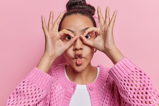 Funny woman makes fish lips and pretends to look through binoculars explores world peers into distance dressed in casual knitted jumper poses against pink background. People fun and curiosity concept
