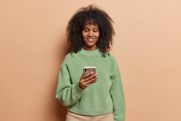 Pretty curly haired woman uses smartphone modern device looks at mobile screen scrolls and enjoys...