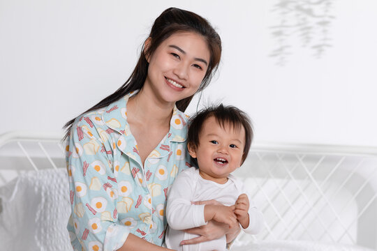 Cute Asian mom hugging her baby boy on bed smile and happiness together. Little baby toddler sit on bed and relax with mother speding time to learning and growth. Good momen of family time. Mother day