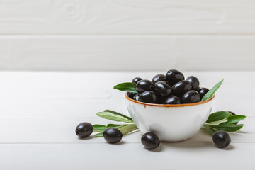 Black olives in a bowl with leaves on a textured white wood background. Delicious and healthy food. Delicacy. Ingredient for salad and cocktails.Mediterranean Kitchen. Close-up. Place for text. 