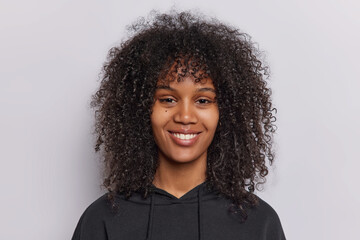 Portrait of happy curly haired teenage girl smiles pleasantly looks directly at camera dressed in casual black sweatshirt isolated over white background beams with joy has genuine smile on face
