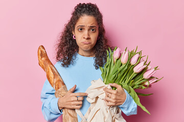 Brazilian woman with dark curly hair poses with bouquet of tulips and french baguette has sad expression being offended wears blue jumper stands indoor against pink background. Holiday preparation