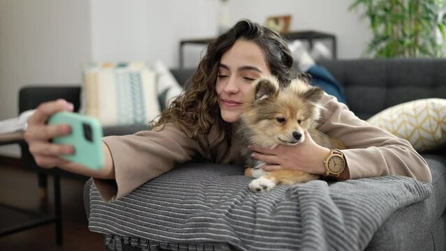 Young hispanic woman with dog taking selfie picture with smartphone lying on sofa at home