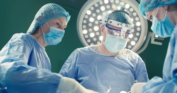 Female intern assists chief doctor and uses scissors. Medical worker dressed up in binoculars. Three doctors perform surgery intervention. Medical workers in special clothes during operation process.