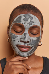 Calm dark skinned woman touches chin gently keeps eyes closed applies facial clay mask gets beauty treatments wears t shirt isolated over brown background. Spa wellness and rejuvenation concept