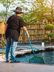 Back view of Asian woman in a black hat and black shirt wearing jeans using dip-net to scoop up fallen leaves and rubbish in koi pond. Cleaning in fish pond. Autumn and summer seasonal pond care work.