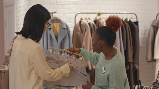 Young positive Black female shop clerk with elegant mini skirt on hanger in hands serving young Caucasian woman doing clothes shopping in modern designer showroom