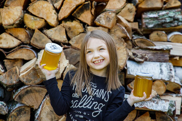 Dziewczynka trzyma słoiki z miodem od pszczół, w tle naturalne drewno, złoty miód pitny - zdrowie, lekarstwo, natura, zdrowy cukier - obrazy, fototapety, plakaty