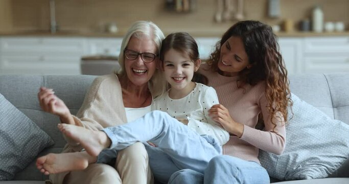 Cheerful grandmother and young mom looking at camera play with little kid on cozy couch in living room, tickle girl, have fun together at home, enjoy carefree weekend. Family ties, playtime activity