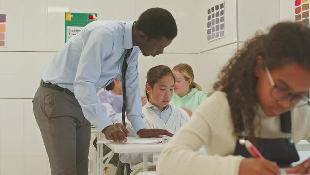 Medium shot of young African American male school teacher checking classwork of multiethnic preteen students sitting at their desks during lesson