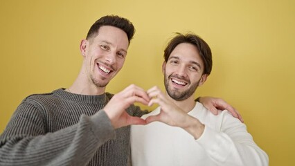 Two men couple hugging each other doing heart gesture over isolated yellow background