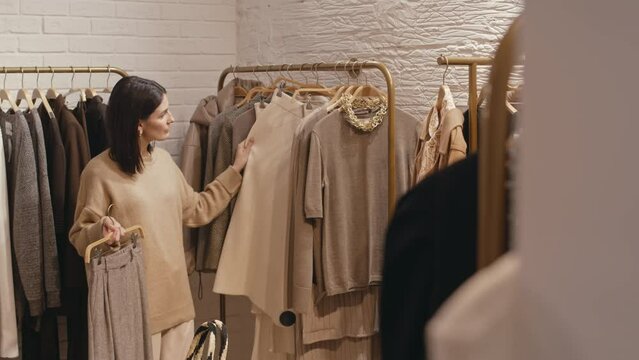 Medium shot of young Caucasian woman choosing beige and camel smart casual outfits hanging on rack in modern designer showroom