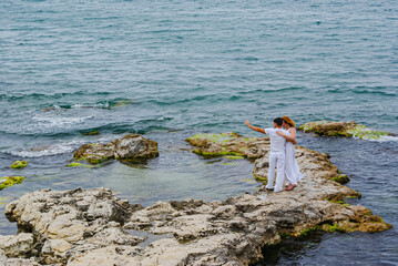 Couple of lovers in white clothes stand on sea rocks and look into distance. Man and woman in white...