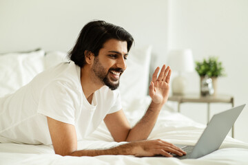 Smiling millennial middle eastern guy woke up, lying in bed, waving hand at laptop webcam, has...