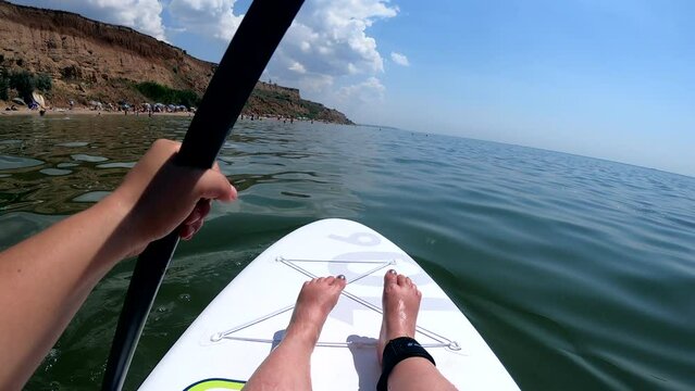 Man floating on SUP board. Person floats on white SUP board and paddles rowing oar on sea surface with waves near shore with blue sky on sunny summer day. Active lifestyle recreation leisure sport POV
