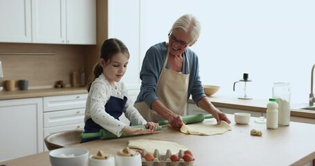 Loving older 60s granny teach her preschooler grandchild to cook, flattening dough for homemade healthy biscuits or pie enjoy talk and time together on weekend at home. Family ties, recipe and hobby