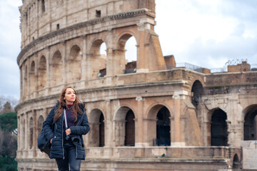 Fototapeta na wymiar Pensive young Latin woman tourist in warm clothes and professional camera standing against Colosseum and looking away while admiring historic architecture during trip through Rome, Italy